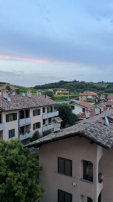 This image shows a peaceful village with tiled-roof houses, balconies, trees, distant green hills, and a soft, pastel sky at dusk, creating a serene atmosphere.