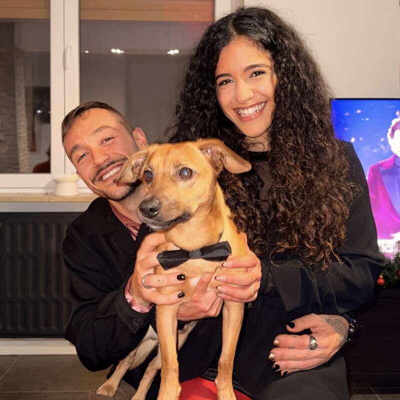 A smiling man and woman pose happily together indoors, holding an adorable brown dog wearing a black bowtie. The cozy atmosphere suggests a warm, joyful moment.