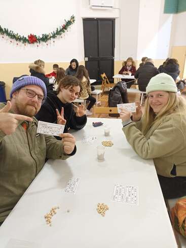 Trois personnes assises à une table jouent au bingo, en souriant et en tenant des cartes. D'autres groupes sont assis derrière eux. Une guirlande festive est accrochée au mur, évoquant une atmosphère joyeuse.