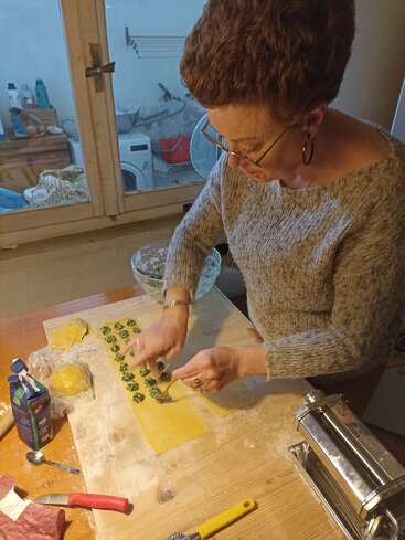 Une femme prépare des pâtes maison, en déposant soigneusement la farce sur des feuilles de pâte. Des ustensiles de cuisine, des ingrédients et une machine à pâtes sont posés sur la table en bois à côté d'elle.
