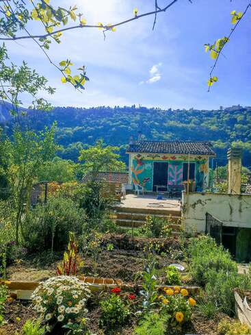 Au premier plan, un jardin éclatant de vie s'épanouit, parsemé de fleurs colorées et d'une végétation luxuriante, qui mène à une petite maison recouverte de fresques, nichée sur le flanc d'une colline boisée baignée de soleil.