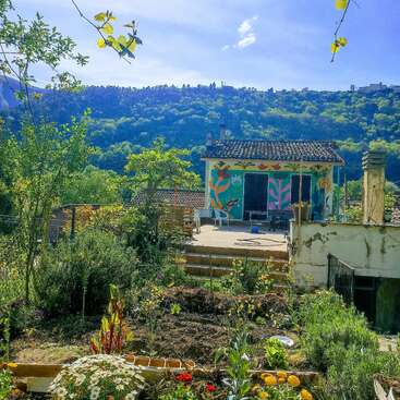 Au premier plan, un jardin éclatant de vie s'épanouit, parsemé de fleurs colorées et d'une végétation luxuriante, qui mène à une petite maison recouverte de fresques, nichée sur le flanc d'une colline boisée baignée de soleil.