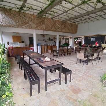 Essa imagem mostra um café ou restaurante espaçoso ao ar livre, com mesas e bancos de madeira, plantas, um design rústico, tabuleiros de xadrez e pessoas reunidas ao fundo.