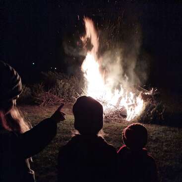 Drei Kinder mit Hüten beobachten nachts ein großes Lagerfeuer im Freien. Die hellen Flammen und der Rauch heben ihre Silhouetten von der dunklen, grasbewachsenen Umgebung ab.