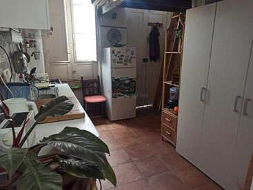 Esta acogedora cocina cuenta con armarios blancos, un frigorífico con imanes, una estantería de madera, una maceta, suelo de baldosas y una ventana que deja pasar la luz.