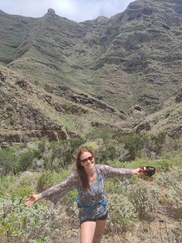 Una mujer sonriente con los brazos extendidos está de pie frente a altas y escarpadas montañas verdes, disfrutando de un día soleado al aire libre, rodeada de arbustos y belleza natural.