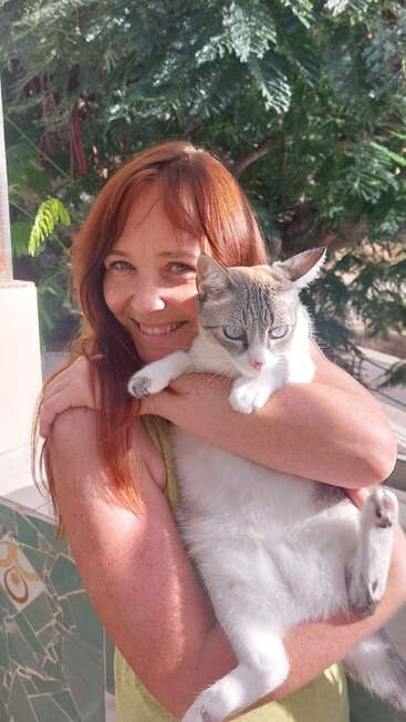 Una mujer sonriente de larga melena pelirroja abraza cariñosamente a un gato blanco y gris. Están al aire libre, rodeados de vegetación, disfrutando juntos de la luz natural del sol.