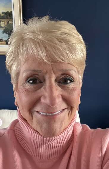 Uma mulher sorridente, de cabelo loiro curto, está vestindo uma blusa de gola alta rosa. Ela está sentada dentro de casa, em frente a uma parede azul-escura com quadros na parede.