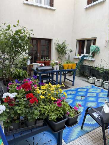 Este pátio aconchegante tem vasos de flores coloridas, plantas verdes, mesas azuis, ferramentas de jardim e um tapete com estampas vivas, criando um refúgio ao ar livre charmoso e colorido.