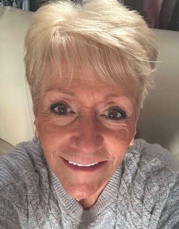 Uma mulher sorridente, com cabelo loiro curto, está sentada em um sofá de cor clara, vestindo um suéter cinza aconchegante. Ela olha diretamente para a câmera, transmitindo calor humano e felicidade.
