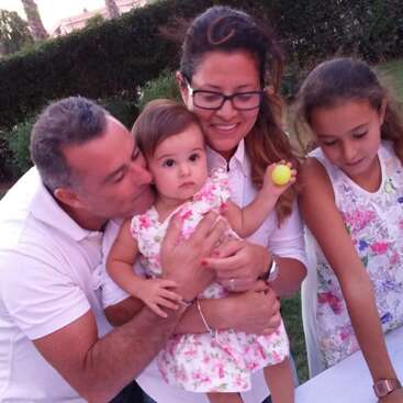 A family of four, including a father, mother, older daughter, and baby girl, share a loving moment outdoors. The baby holds a yellow ball.