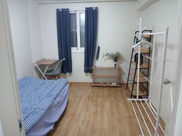 This room features a neatly made bed, study desk, chair, window with blue curtains, shelves, a small table with decor, and a clothing rack. Minimalist style.