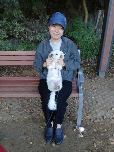 A smiling person wearing a blue cap sits on a wooden bench, holding a small white dog upright, surrounded by greenery and fallen leaves outdoors.