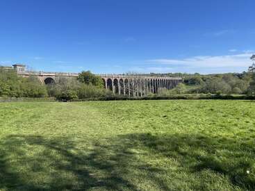 Ein langes, historisches Backsteinviadukt erstreckt sich über eine üppig grüne Wiese, umgeben von Bäumen und unter einem strahlend blauen Himmel an einem strahlend sonnigen Tag.