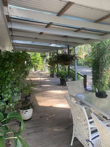 A covered wooden patio featuring white wicker chairs and a glass table, surrounded by lush green potted plants and hanging baskets, creating a relaxing outdoor space.