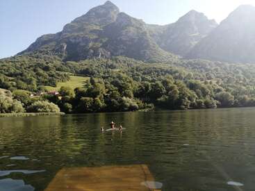 Uma pessoa nada tranquilamente em um lago calmo cercado por florestas verdejantes. Ao fundo, montanhas imponentes e escarpadas se erguem dramaticamente sob um céu claro.