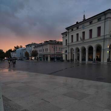 The image depicts a serene city square at dusk, featuring a large white building with arches and columns, surrounded by people and trees under a cloudy sky.