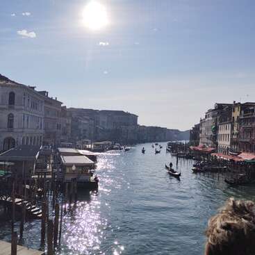 The image depicts a serene canal scene in Venice, Italy, with gondolas and buildings lining the waterway, bathed in sunlight and surrounded by a tranquil atmosphere.