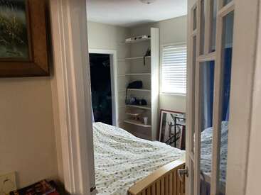 Ein gemütliches Schlafzimmer mit einer gemusterten Tagesdecke, einem offenen Kleiderschrank, Einbauregalen, gerahmten Kunstwerken, einem Fenster mit Jalousien und einem teilweise sichtbaren Bettgestell aus Holz.