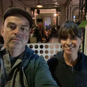 Um homem e uma mulher tiram uma selfie em um restaurante ou café com pouca luz, enquanto outras pessoas estão sentadas no fundo, num ambiente aconchegante e bem iluminado.