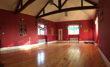 A imagem mostra uma sala espaçosa com teto alto, com uma grande janela e piso de madeira. As paredes são pintadas de branco e a sala é bem iluminada. A atmosfera geral é clara e arejada.