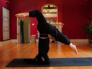 Uma pessoa pratica ioga dentro de casa, equilibrando-se na cabeça e nos antebraços, na postura da postura sobre a cabeça. A sala tem paredes vermelhas, piso de madeira e iluminação aconchegante.