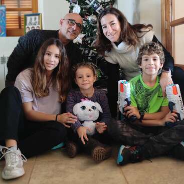 The image depicts a family of five posing for a photo in front of a Christmas tree, with two children holding Nerf guns and a stuffed animal.