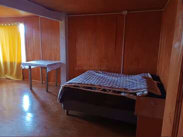 Dieses Zimmer hat hölzerne Wände und Böden, ein Bett mit einer Steppdecke, einen kleinen Nachttisch, einen Tisch mit einem Tuch und gelbe Vorhänge vor dem Fenster.