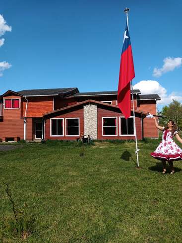 Eine Frau in einem traditionellen Kleid tanzt an einem sonnigen, klaren Tag fröhlich neben einer großen chilenischen Flagge vor einem rustikalen roten Haus.