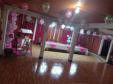 Dieses Bild zeigt einen geschmückten Partyraum mit rosafarbenen und weißen Luftballons, langen Tischen mit rosafarbenen Tischdecken, Stühlen, Holzboden und festlichen Luftschlangen, die von der Decke hängen.