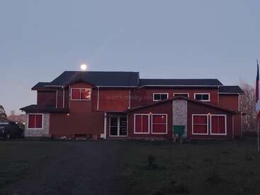 Ein großes zweistöckiges Haus mit roten Wänden und steinernen Akzenten steht in der Abenddämmerung, beleuchtet vom Vollmond über dem Kopf und warmen Lichtern im Inneren.