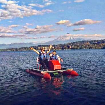 Two people wearing life jackets enjoy a pedal boat ride on a tranquil lake, with a snow-capped volcano and a scenic landscape in the background.