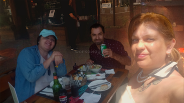 Tres personas están sentadas a la mesa de un restaurante al aire libre, sonriendo a la cámara. En la mesa se ven bebidas, comida y pan. El ambiente parece relajado y amistoso.