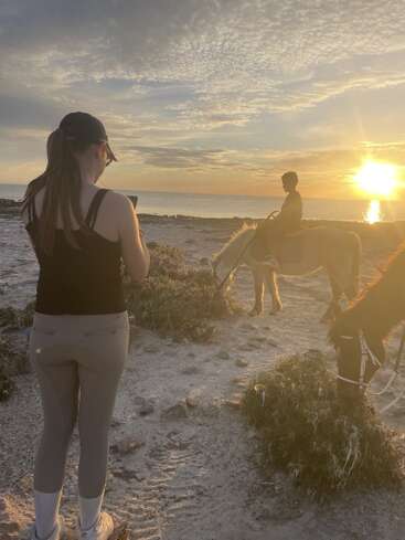 Une femme se tient sur une plage de sable et de rochers et prend une photo d'une personne à cheval au coucher du soleil, dans une lumière magnifique, avec des nuages et un paysage paisible.