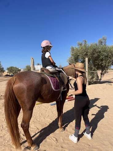 Une jeune fille coiffée d'un casque rose est assise sur un cheval brun. Un moniteur se tient à ses côtés et la guide dans un espace extérieur sablé, sous un ciel bleu limpide.