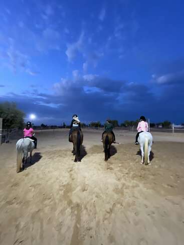 Au crépuscule, quatre personnes montent à cheval dans un manège de sable, sous un ciel d'un bleu éclatant parsemé de nuages. Les cavaliers portent des casques et montent chacun un cheval d'une couleur différente.