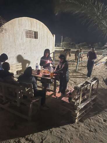 Un groupe de personnes est réuni autour d'une table en bois à l'extérieur, de nuit, dans la douce lueur des lampes. À l'arrière-plan, deux chevaux se tiennent derrière une clôture.