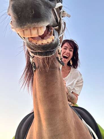Une femme rayonnante est à cheval ; tous deux sourient largement à la caméra. La photo est prise en contre-plongée, capturant leurs rires sur fond de ciel dégagé.