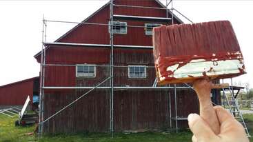 Une personne peint au pinceau un grand bâtiment, probablement une grange ou un hangar, avec un grand terrain et une clôture en arrière-plan. Le bâtiment est blanc avec un toit vert.
