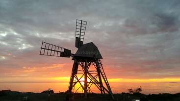 L'image représente un moulin à vent au corps blanc et aux pales noires, situé dans une zone herbeuse, entouré d'une clôture, avec un petit bâtiment à l'arrière-plan.