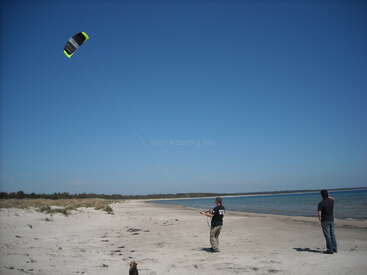 L'image représente deux hommes sur une plage, l'un faisant voler un cerf-volant avec une voile noire et jaune, tandis que l'autre observe, sur fond de ciel bleu clair.