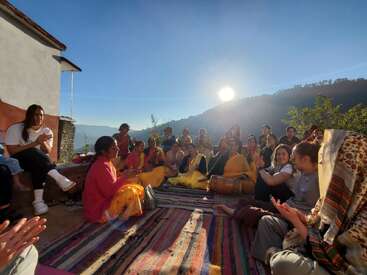 Eine Gruppe von Menschen sitzt im Kreis auf bunten Teppichen im Freien, teilt sich und klatscht. Die Sonne geht über den fernen Hügeln auf und schafft eine heitere und fröhliche Atmosphäre.