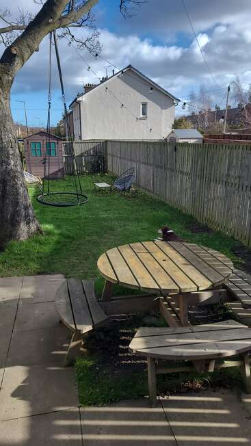 A fenced backyard with green grass, a tree swing, round wooden picnic table, small shed, outdoor chairs, fairy lights, and a dog lying peacefully.