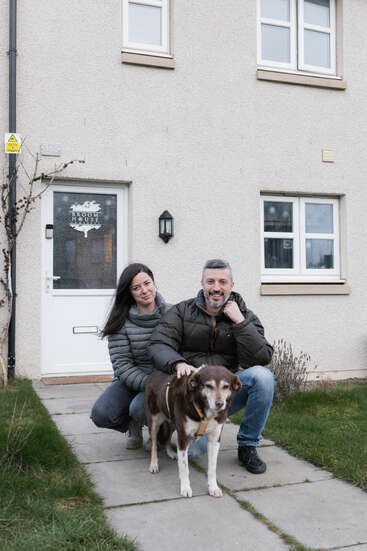Ein lächelndes Paar kniet vor einem beigen Haus, an dem ein Schild mit der Aufschrift „Broom House“ hängt. Sie streicheln einen braun-weißen Hund auf dem Weg.