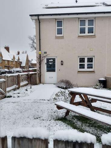 Ein schneebedeckter Garten mit einem hölzernen Picknicktisch, einem hellen Haus mit weißen Fenstern und einem Weg, der zur Haustür führt. Alles wirkt friedlich und ruhig.