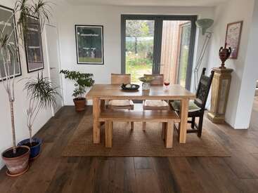 Uma área de jantar aconchegante com uma mesa de madeira, cadeiras variadas, plantas de interior, pôsteres, uma luminária de chão e vista para o jardim através de portas de vidro, criando uma atmosfera natural e convidativa.