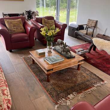 Uma sala de estar aconchegante com poltronas vermelhas, mesa de centro rústica, lírios frescos, escultura com tema de caça, tapetes estampados, móveis vintage, janelas grandes e vistas tranquilas para o jardim.