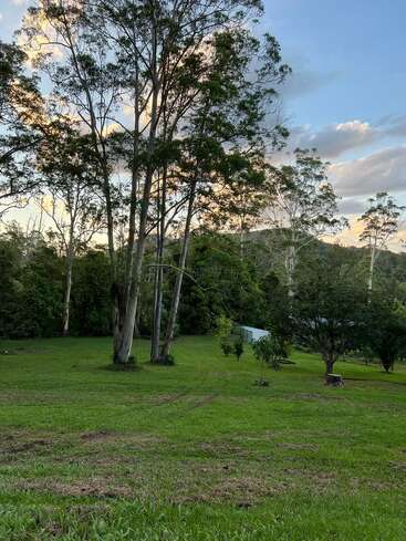 The image depicts a serene landscape featuring a grassy field with trees, a small white building, and a distant mountain range under a blue sky with clouds.