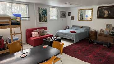 This cozy studio apartment features a bunk bed, sofa, and bed, adorned with art, mirrors, red rug, table, chairs, and windows providing natural light.