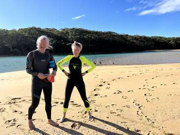 Two people in wetsuits stand barefoot on a sandy beach with footprints, near calm water, under a blue sky; pelicans relax by the shoreline behind them.